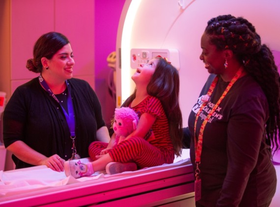 Little girl on MRI with two female providers smiling next to her